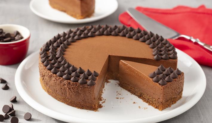 Fotografia em tons de branco e vermelho de uma bancada branca com um prato branco, sobre ele uma torta de chocolate. Ao lado um paninho vermelho com uma espátula. Ao fundo um prato branco com uma fatia de torta e um potinho com gotas de chocolate.