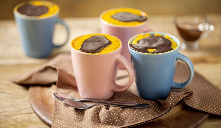 Canecas nas cores azul e rosa comporta o bolo de cenoura. As canecas estão em cima de uma mesa de madeira e um pano marrom.