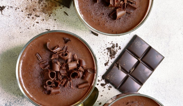 Fotografia vista de cima, em tons claros, de três recipientes de vidro sobre uma mesa clara. Dentro dos potinhos tem a sobremesa, finalizada com ganache e raspas de chocolate, e ao redor tem raspas e algumas barras pequenas de chocolate.