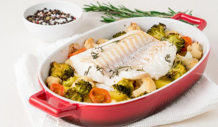 fotografia em tons de branco, vermelho e amarelo de uma bancada branca vista de cima. Contém um recipiente oval vermelho, com um pedaço de bacalhau e legumes em volta.