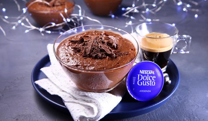 Fotografia em tons de azul em uma bancada de cimento queimado, um prato redondo azul com uma taça de vidro com a mousse de chocolate dentro e um paninho branco ao lado. Ao lado, uma xícara de vidro com café e atrás decoração com luzes natalina.