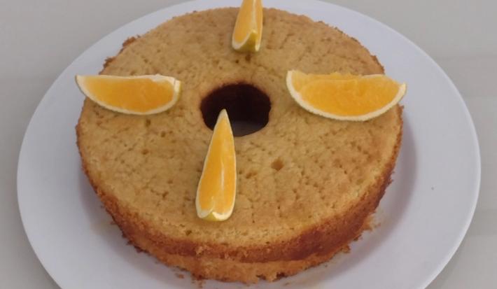 Fotografia de um bolo de laranja feito na forma de fundo central, sobre um prato de vidro branco. Sobre o bolo tem quatro pedaços de laranja cortados.