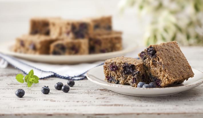 Fotografia em tons de azul, marrom e branco, tendo à frente prato branco com pedaços de bolo com blueberry e blueberries espalhadas na bancada, ao fundo outro prato com pedaços de bolo sobre guardanapo branco de borda azul, tudo sobre bancada branca.