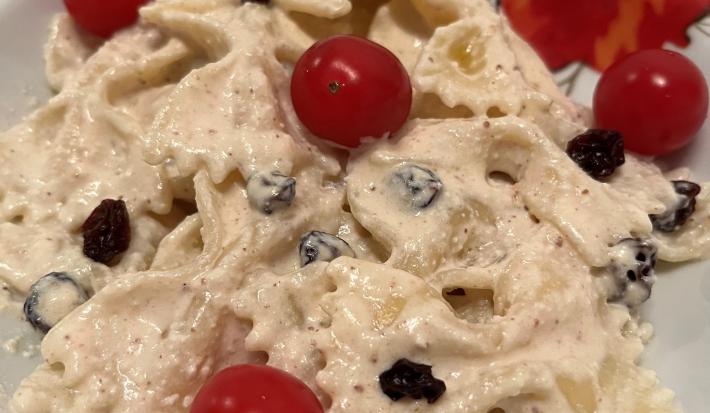 Fotografia de uma masssa farfalle cremosa com uvas passa e tomates cereja dentro de um prato branco de vidro.