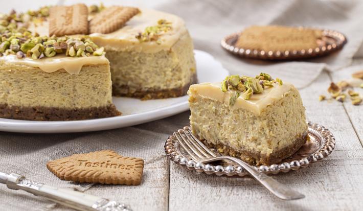 Foto da receita de Cheesecake de Pistache. Observa-se a torta com um pedaço cortado em um prato de sobremesa, decorado com pistaches e uma calda na cor branca.