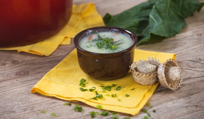 Imagem de um bowl marrom com a receita de caldo verde sobre um pano amarelo. Ao fundo uma panela vermelha e uma folha de couve.