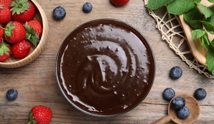 Fotografia vista de cima de um recipiente redondo com a sobremesa, finalizada com ganache, ao lado de um potinho de madeira com morangos inteiros, sobre uma mesa de madeira. Ao redor dos recipientes em mirtilos e mais morangos inteiros, espalhados.