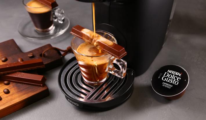Fotografia em tons de preto em uma bancada de madeira cinza, uma máquina de Dolce Gusto com uma xícara de vidro e o café dentro dela, por cima, um KitKat sendo derretido pelo café caindo da máquina. Ao lado, tábua com gotas de chocolate.