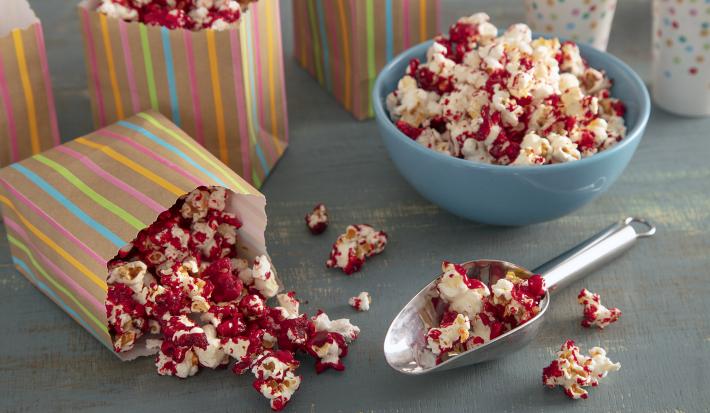 Foto da receita pipoca doce vermelha. Observa-se uma tigela azul com a pipoca e, ao lado, saquinhos estampados com a pipoca também.