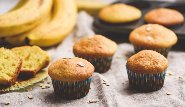 Fotografia de quatro muffins de banana e aveia dentro de uma forminha de papel colorida sobre uma toalha de mesa em tons de bege. Ao lado, um cacho de bananas, duas fatias do bolo, e ao fundo, dois muffins em uma forma preta de bolinhos.