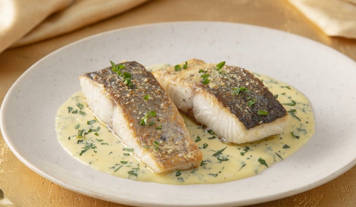 Filé de peixe branco dourado servido no molho cremoso de ervas, apresentado em prato fundo branco com acabamento sofisticado.