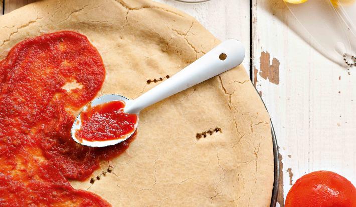 Fotografia em tons de vermelho e branco, de uma bancada de madeira com uma massa redonda de pizza, com molho de tomate pela metade e uma colher prateada. Ao redor potinhos, ovos, tomates e azeite.