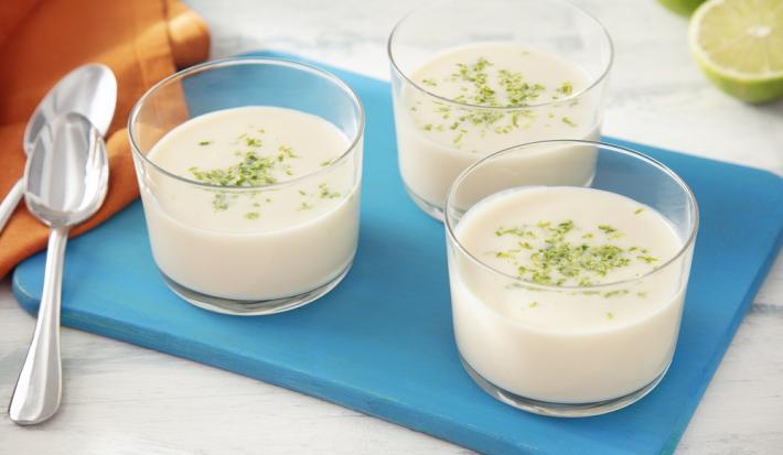 Foto da receita de mousse de limão zero lactose servida em três porções em taças sobre uma tábua azul com um pano laranja ao lado além de duas colheres prateadas