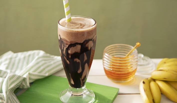 Fotografia em tons de marrom em uma bancada de madeira de cor branca. Ao centro, uma tábua de madeira de cor verde contendo uma taça de vidro com o smoothie. Ao lado, há um pano branco listrado e ao fundo, um pote de mel e um cacho de banana.