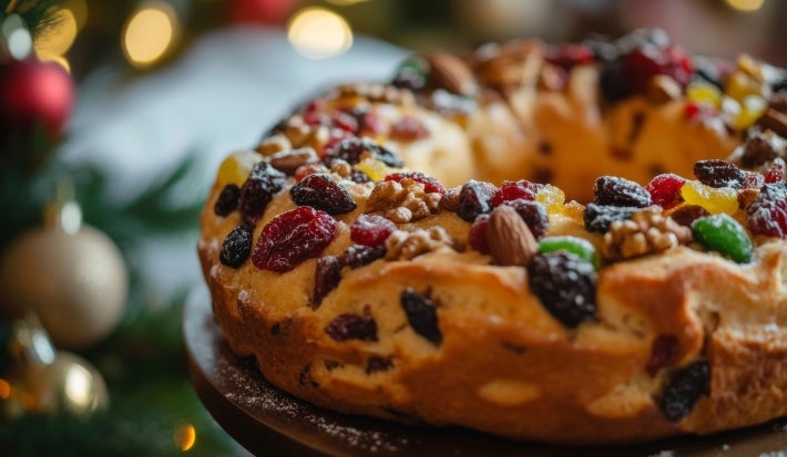 Fotografia de um bolo de frutas tradicional, adornado com frutas secas e nozes, levemente polvilhado com açúcar de confeiteiro, em um ambiente natalino com uma árvore de Natal ao fundo.