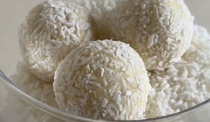 Fotografia em tons claros de várias trufas de chocolate branco passadas no coco ralado dentro de um recipiente de vidro fundo sobre uma mesa branca.