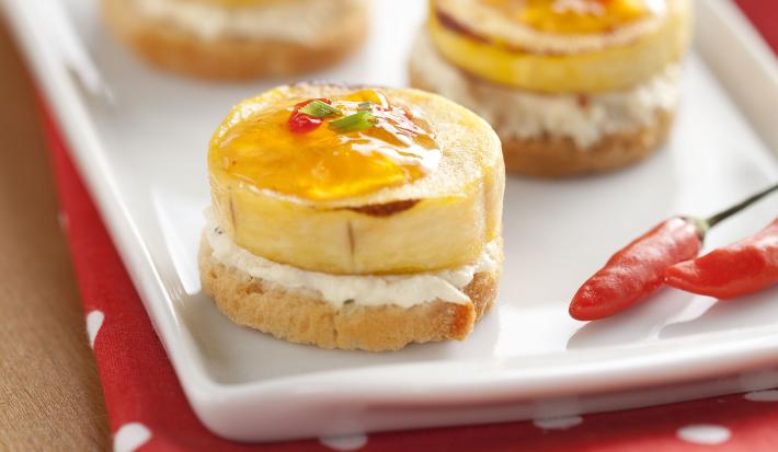 Fotografia em tons de vermelho em uma bancada de madeira, uma toalha vermelha com bolinhas brancas, um recipiente branco retangular com os canapés de banana com geleia de pimenta.