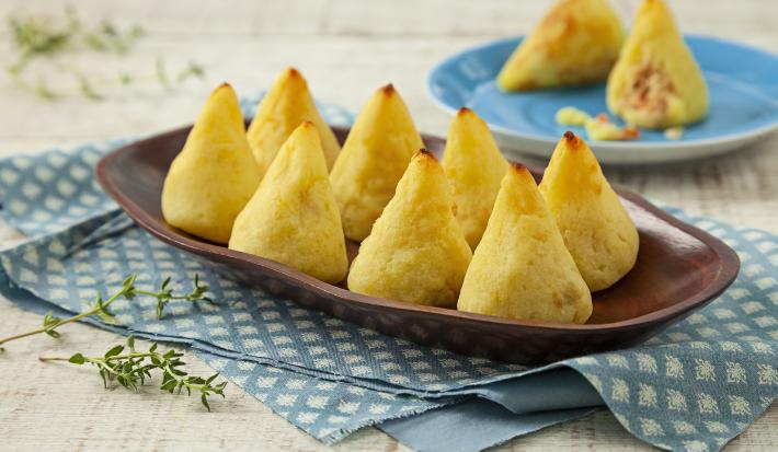 Fotografia em tons de azul e amarelo em uma bancada de madeira clara, um paninho azul listrado, um recipiente oval marrom com coxinhas. Ao fundo, um prato azul redondo fundo com duas coxinhas em cima dele, uma delas está aberta.
