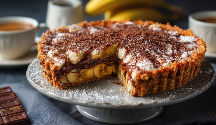 Em cima de uma mesa, banoffee com massa de biscoitos crocante com uma fatia cortada. Dentro da banoffee, bananas em rodelas, doce de leite, decorado com chantilly e canela em pó.