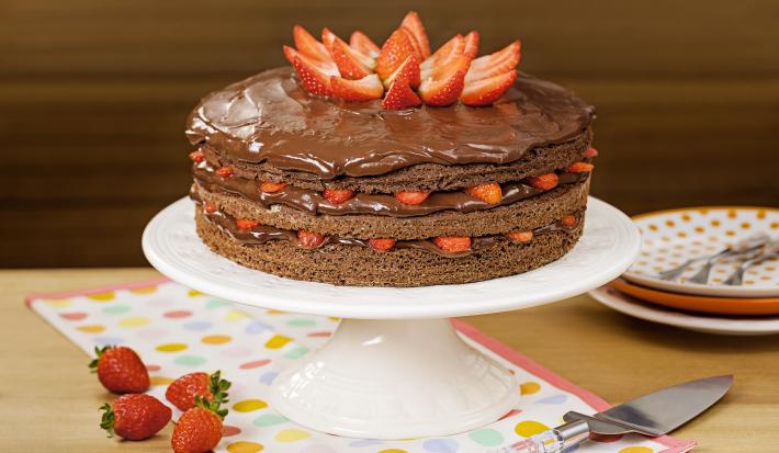 Fotografia em tons de marrom e vermelho de uma bancada de madeira, uma boleira com o bolo de chocolate e morangos. Ao lado quatro morangos para decorar, um espátula para servir e três pratos coloridos em tons de laranja com garfos.