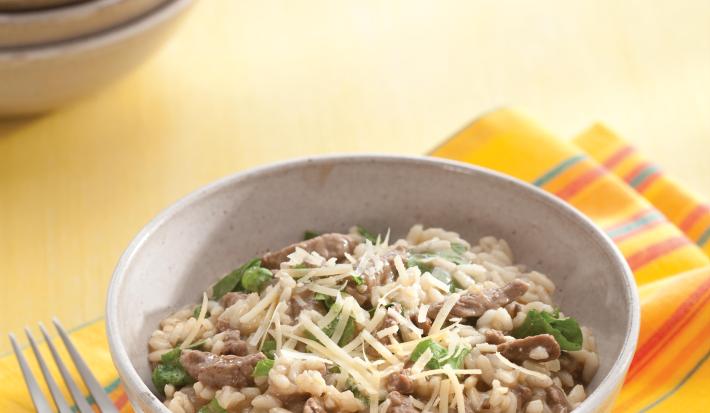 fotografia em tons de bege, cinza e amarelo de uma bancada bege vista de cima. Contém um pano amarelo com um recipiente redondo cinza com o risoto, com pedaços de carne e ao lado um garfo para servir.