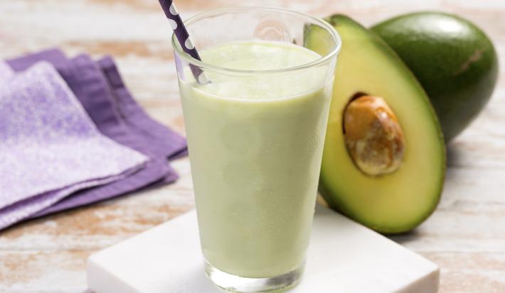 fotografia em tons de bege, verde e roxo de uma bancada bege vista de frente. Contém um copo transparente com a vitamina de abacate, ao fundo do lado direito uma fatia de abacate e do lado esquerdo um pano roxo