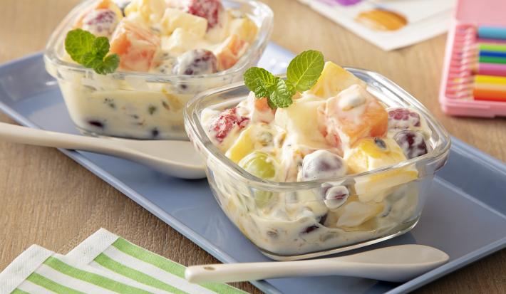 Fotografia de dois recipientes quadrados pequenos de vidro com salada de frutas e creme de maracujá com ninho em cada um, misturado. Por cima deles tem algumas folhas de hortelã, ao lado tem uma colher clara, também sobre um apoio retangular azul.