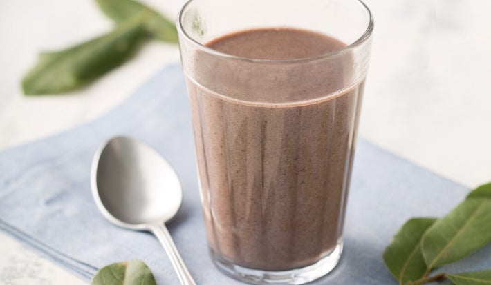 Foto de um copo de vidro com feijoada cremosa de textura lisa e cor marrom-escura. O copo está sobre um guardanapo azul claro, ao lado de uma colher prateada. Folhas verdes decoram a cena, que tem fundo claro e iluminação suave