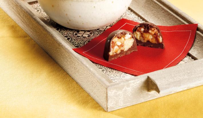 Fotografia em tons de branco, vermelho e marrom. Contém um pano vermelho com um bombom cortado ao meio e ao lado um recipiente redondo branco com vários bombons