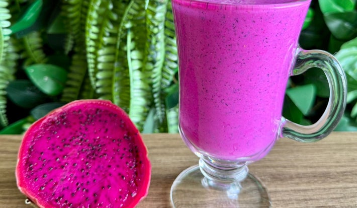 Fotografia mostra um copo com bebida rosa, ao lado metade de uma pitaya aberta.