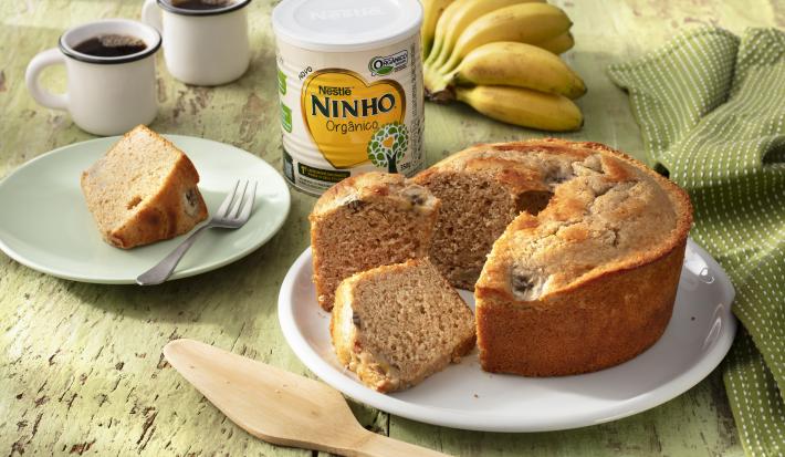 Fotografia em tons de verde de uma bancada verde com um prato branco redondo, sobre ele um bolo de banana. Ao lado um prato com uma fatia de bolo e um garfo, duas xícaras de café, um paninho verde com um cacho de banana e uma lata de NINHO Orgânico