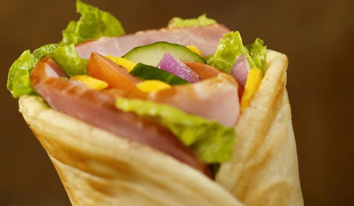 Fotografia de um lanche fechado com pão sírio, alface, tomate, pepino, cebola, molho e peito de peru, e um fundo escuro.