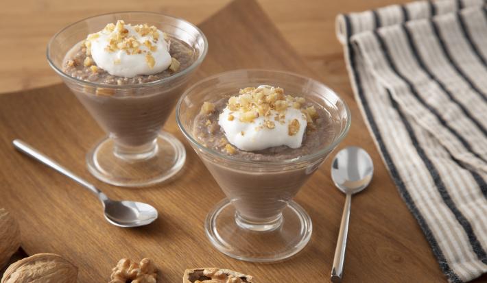 Foto da Receita de Creme de Nozes. Observa-se dois potinhos de vidro transparentes com o creme de nozes dentro. Ao lado dos potinhos, colherinhas prateadas e um guardanapo listrado ambientam a foto.