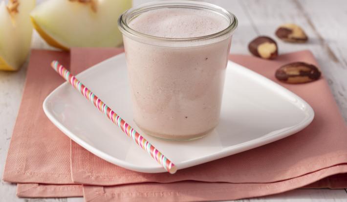 Fotografia em tons de branco e rosa de uma bancada branca com um paninho rosa e um prato quadrado branco, sobre ele um copo de vidro com o Smoothie e um canudo listrado rosa. Ao fundo duas fatias de melão e castanhas-do-pará.