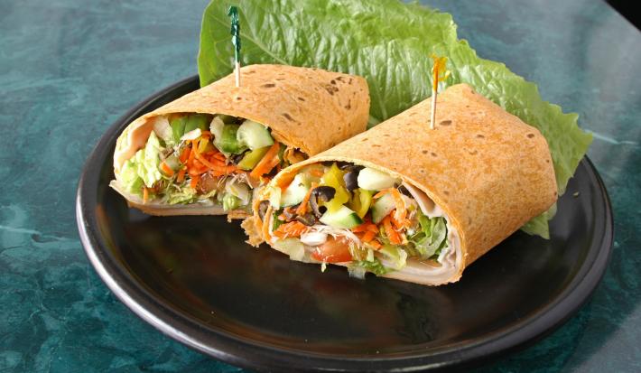 Fotografia de wrap de cebolinha no pão sírio cortado pela metade, com tomate, pepino e alface. O wrap está em um prato preto sobre uma mesa de mármore em um tom de verde.