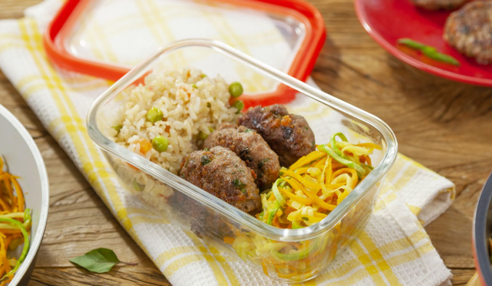 Fotografia em tons de amarelo em uma mesa de madeira com um pano de prato branco com listras amarelas, um potinho de marmita de vidro com o arroz integral, o mini hambúrguer e o macarrão de legumes