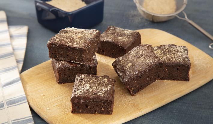 Foto da receita de Brownie com Farinha Láctea NESTLÉ. Observa-se uma tabua de madeira com 6 brownies quadrados com farinha láctea polvilhada