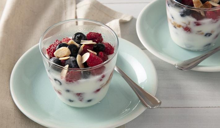 Foto em tons de azul claro da receita de parfait de iogurte com frutas vermelhas servida em dois copos altos sobre pratos pequenos azuis