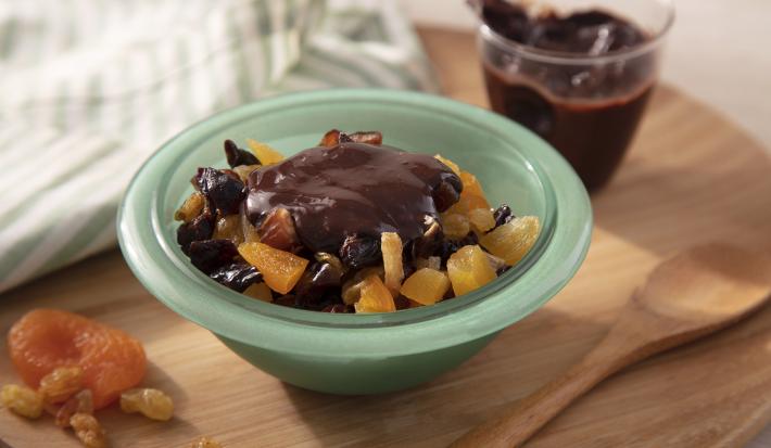 Fotografia em tons de amarelo em uma bancada de madeira de cor marrom. Ao centro, um recipiente verde contendo a salada de frutas. Ao lado, há algumas frutas picadas com uma colher de madeira ao lado. Ao fundo, um pote com a ganache de chocolate.