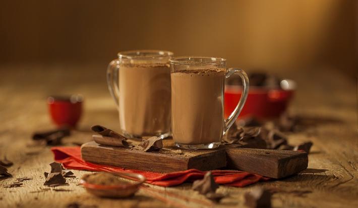 Fotografia em tons de marrom em uma bancada de madeira de cor marrom. Ao centro, um pano vermelho contendo 2 xícaras com o chocolate quente e ao redor há alguns pedações de chocolates espalhados.