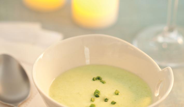 Fotografia em tons de verde em uma mesa de madeira com uma toalha verde, ao centro um potinho branco redondo fundo com o creme de abobrinha com cebolinha decorando. Ao fundo, duas velas acesas e uma taça de vidro com água.