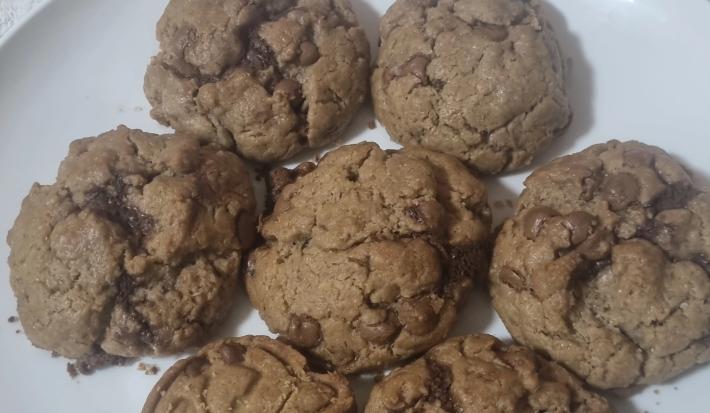 Fotografia de sete cookies grandes com gotas de chocolate sobre um prato grande e raso, de cor branco.