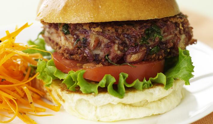 Fotografia de um hambúrguer com carne bovina, salada, ovo e queijo, acompanhado de cenoura cozida. O hambúrguer está sobre um prato branco que está apoiado em um pano cor de salmão. Tudo está sobre uma mesa verde claro.