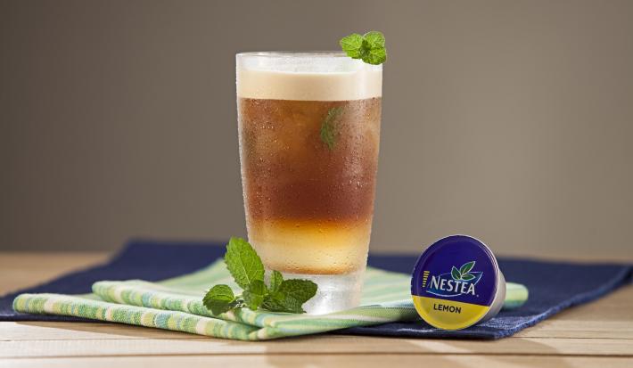 fotografia em tons de marrom e roxo de uma bancada marrom vista de frente. Contém um pano roxo e um pano em tons de verde e branco com um copo transparente que contém a bebida de nestea Limão com folhas de hortelã na borda do copo