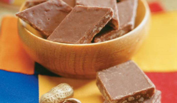 fotografia em tons de marrom, azul e amarelo. Contém um pote redondo marrom com pedaços quadrados de amendoim com chocolate.
