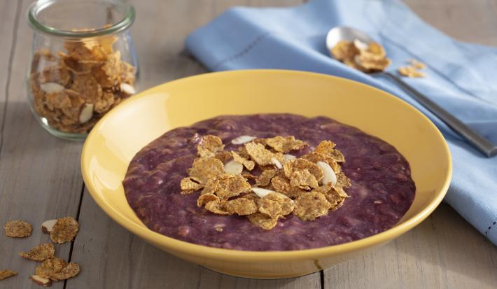 Fotografia em tons de roxo em uma bancada de madeira escura com um pano azul claro ao lado, um prato redondo fundo amarelo com o creme de açaí dentro dele e decorado com cereais. Ao fundo, um potinho de vidro com cereais.