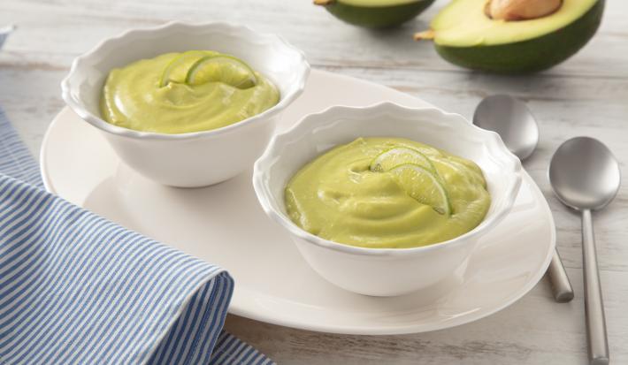 Fotografia em tons de branco e verde de uma bancada, ao centro dois potinhos com creme verde de abacate. Ao fundo, um abacate cortado ao meio.