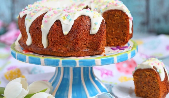 Fotografia de um bolo bundt decorado, coberto com um generoso glacê branco que escorre pelas laterais, adornado com confeitos coloridos. O bolo está em um suporte listrado. Ao lado do suporte, prato branco com uma fatia do bolo fofino e dourado.