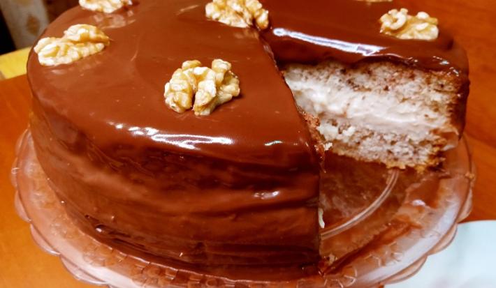 Foto em tons de marrom da receita de bolo de nozes festivo servida em uma porção grande com uma fatia retirada, mostrando o interior do bolo com o recheio. Está sobre uma bailarina (base utilizada na cozinha) em cima de uma mesa de madeira