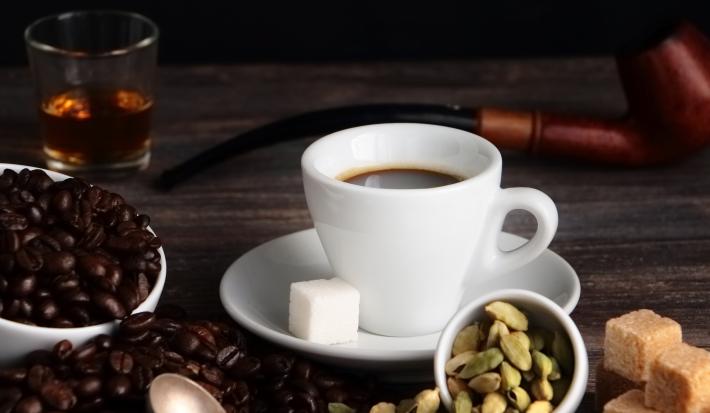 Fotografia em tons de marrom com uma xícara branca ao centro. Dentro da xícara existe um café acompanhado com um quadrado de açúcar. Ao lado existe uma vasilha com grãos de café.